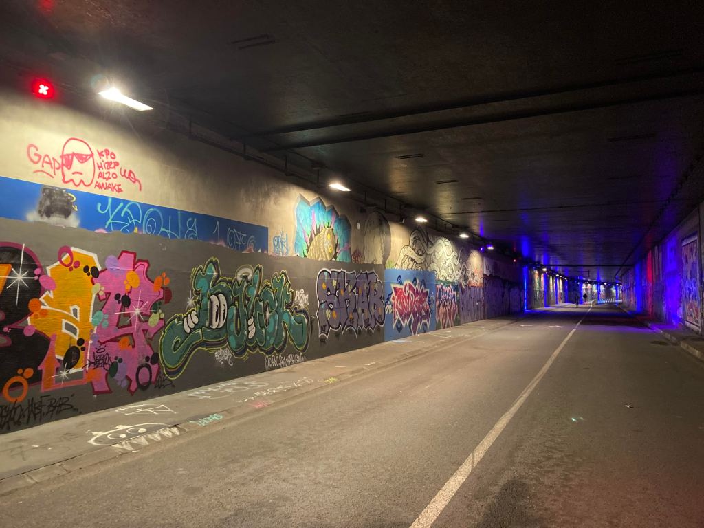 The start of a concrete underground tunnel, showing colourful graffiti tags in the foreground and neon blue spotlights in the background